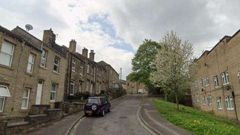 A residential street which slopes uphill. Terraced houses are on both sides of the road and a parked car is outside one of the homes. Trees are also visible.