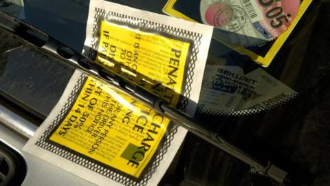 A close up of a parking charge notice is under the windscreen wiper of a car.