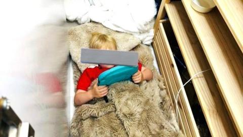 Young child with face hidden (by grey bar) under covers inside a cupboard.