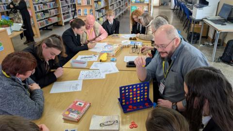 Adults and children sit around a table in a library playing chess, the table-top game Connect 4 and doing word searches.