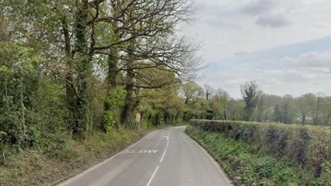 A Google Maps image of the A287 Folly Hill in Farnham. There is a two-lane road with trees on one side a green bushes on the other. 