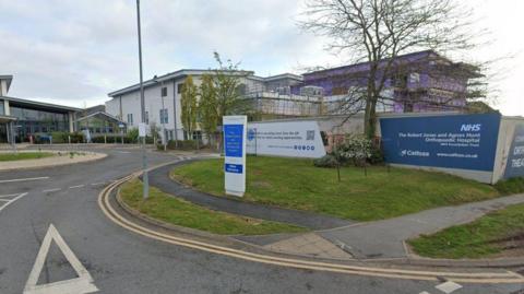 The front entrance of a hospital with large white buildings and a small roundabout in front of a glass entrance building. There is a blue and white sign at the front that reads "The Robert Jones and Agnes Hunt Orthopaedic Hospital"