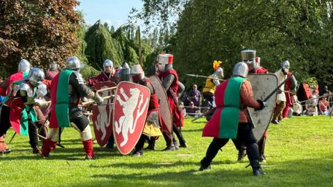 A crowded medieval battle reenactment scene, with participants in armour and helmets pushing forward with shields and long wooden spears, simulating close combat.
