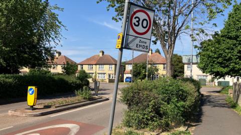 A view of a road junction with a traffic island in the middle and speed restriction signage on the road at at the side.