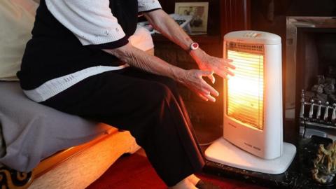 The hands of an elderly woman, who is sitting in a lounge, are stretched towards an portable electric heater. She is wearing red slippers.