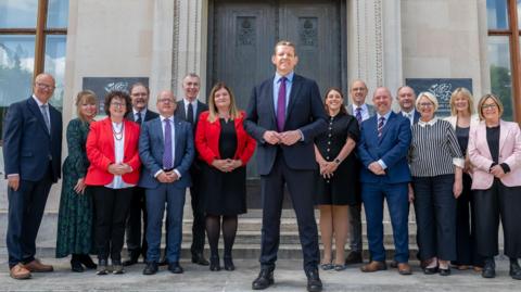 The first minister and cabinet ministers stand on steps in front of a grey building with a brown door.