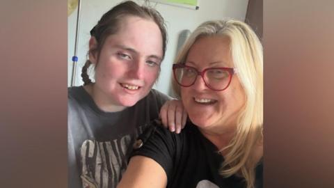 A young woman with long plaited brown hair and wearing a Guns N' Roses T-shirt is smiling. Her hand is resting on the shoulder of her mum, who has long blonde hair, is wearing a dark T-shirt and is also smiling. They are in a hospital room.