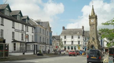 A main street in Machynlleth leading to a tall stone clock tower in a square and townhouses around the edges. There are cars on the road. It is a blue sky with some clouds.