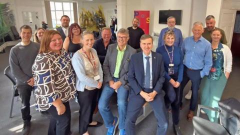 Members of the board standing in a semi-circle around a room with Andrew Pakes in the middle, leaning against a table.