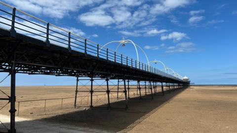A picture of Southport Pier, taken from the sandy beach below. The pier stretches out to the sea on a sunny day with bright blue skies and a handful of white clouds.