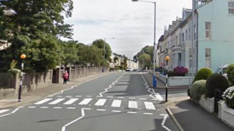 The white striped lines of a pedestrian across Woodbourne Road, with a cement bus shelter on the left and houses on the right.