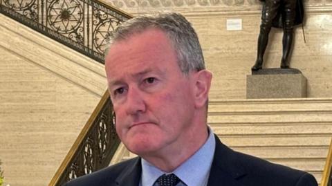 Conor Murphy in the Great Hall at Stormont. He is wearing a navy blazer, light blue shirt and polka dot tie. Marble staircase is in the background.