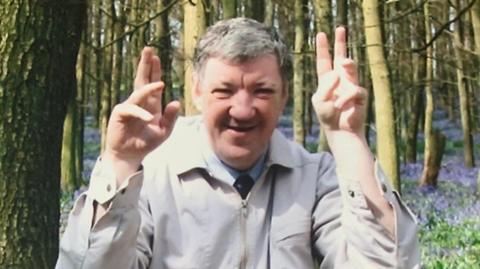 A man wearing a coat and shirt and tie smiling with his arms in the air in a woodland with bluebells.