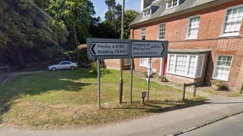 Road signs pointing both left and right on a grass verge with a brick building to the right.