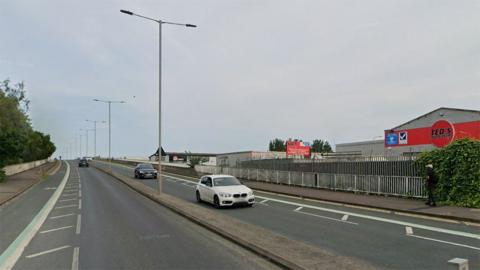 Hessle Road flyover with cars driving over it and a man walking down the right hand pavement. It has on-road cycle lanes and street lights along the central reservation. There are warehouses in the background.