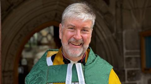 The Dean of Guernsey the Very Reverend Tim Barker after his final Liberation Day service in May. He is smiling straight at the camera and wearing church robes. He is stood outside the church. 