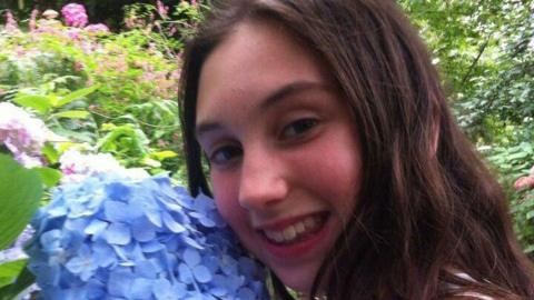 A young girl with rosy cheeks smiles widely with her face rested on a blue hydrangea.