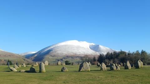 A snow-topped mountain with the stone circle in the foreground which is made up of around 20 large stones on a grassy field. There are fields and trees below the mountain. There are no clouds in the sky and it is a bright, light blue.