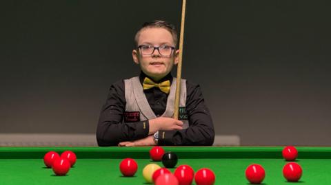 A young boy with glasses is wearing a black shirt, yellow bow ties and tweed waistcoat as he holds a snooker cue with his arms crossed on a snooker table with scattered balls