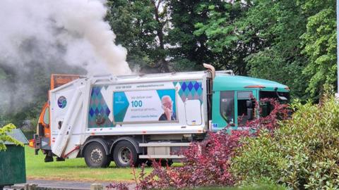 A side view of a white bin lorry, with a turquoise cab. White smoke is billowing from the top of the lorry 