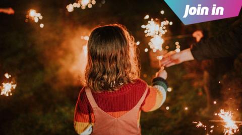 Child is seen holding a sparkler with an adult's hand also in shot helping her keep it upright. She is facing away from the camera standing on a grass surface.