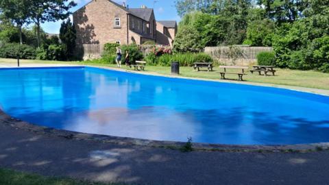 A large blue paddling pool set in parkland. A pavement surrounds one side, and grass on the other, with picnic benches.