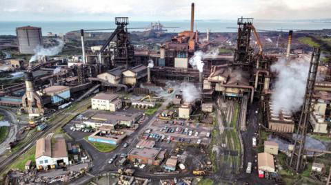 An aerial shot of Tata Steel's Port Talbot steelworks in south Wales. Smoke is coming out of a number of chimneys across the site, while cars are parked in the middle. To the right-hand side rail tracks lead out the works, while the sea can be seen behind it. 