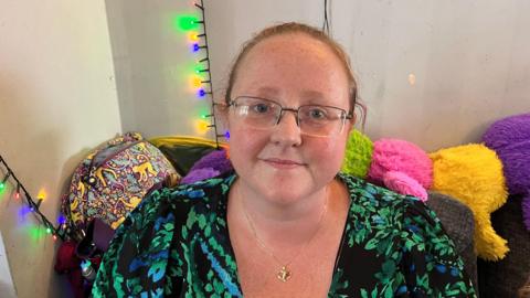 Amy Landsbury sits on a sofa wearing glasses and a green and blue top with bright coloured cushions behind her. 