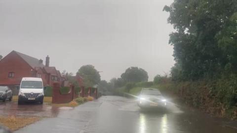 A flooded road with a car driving slowly through the water. There are houses on the left, hedge on the right.