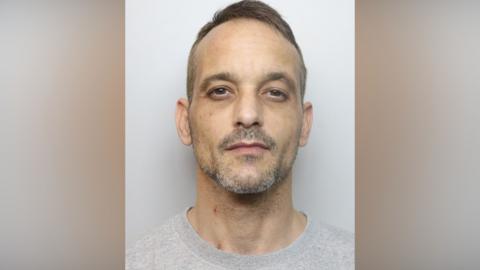 A man in a grey jumper stands looking at the camera in a police mug shot.