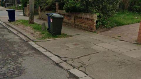 A residential street in Zetland. The pavement has large cracks in the concrete footpath path which looks uneven. There are wheelie bins and grass on part of the pavement, which is next to a road.
