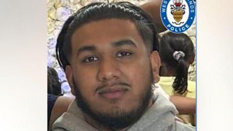 Riyan Ali is wearing a grey hoodie. He has long dark hair pushed back by a hairband, and has a black moustache and beard