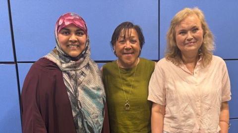 Three people standing side by side in an indoor room with blue wall panels, posing for a photo. The person on the left wears a long maroon outfit with a patterned headscarf, the person in the middle wears a green top and trousers with a necklace, and the person on the right wears a light pink short-sleeved blouse with a long white skirt. All three face the camera against a clean, modern office-style background.
