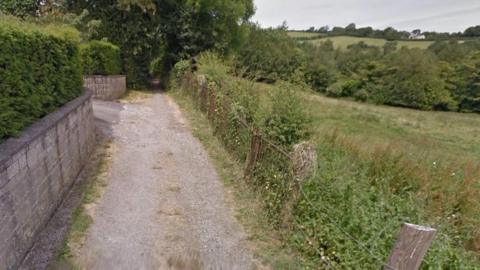 A gravel country lane with fields on the right hand side and a grey brick wall and a hedge on the left. Trees crowd over the path. 