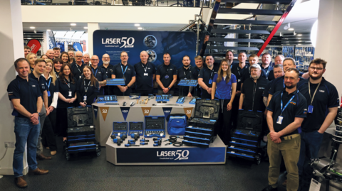 A group shot of about two dozen male and female employees starting to mark the anniversary. They are stood in a blue uniform around some of their products.