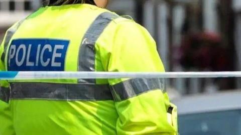 The back of a police officer, wearing a yellow hi-vis jacket with "police" on the back, with blue and white police tape behind him.