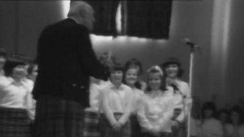 A black and white photo shows a school choir dressed in white shirts and kilts singing, the choir master stands facing the choir and with his back to the camera