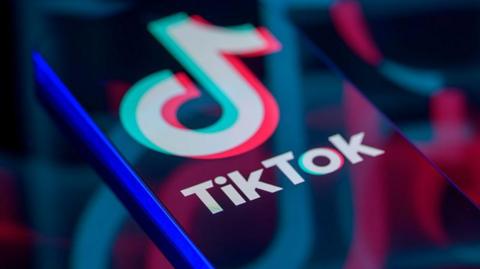 TikTok logo displayed on a smartphone screen, with enlarged, faded versions of the logo reflected across it.