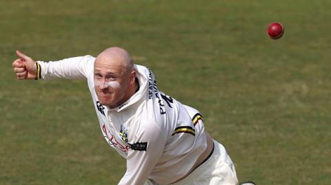 Left-arm spinner Callum Parkinson bowling a ball for Durham with the red cricket ball in the top right corner of the photo
