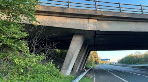 A view of a concrete motorway bridge next to a three lane motorway carriageway. A blue motorway lane sign is pictured in the distance.