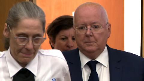 Man in suit and glasses with balding hair flanked by two female security guards.