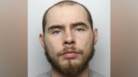 A headshot of a man wearing a grey t-shirt. He has blue eyes, a close-shaven head and a brown beard.
