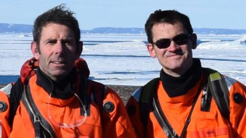 Prof Dave Roberts and Prof Colm O'Cofaigh in bright orange gear and looking at the camera. They are standing in front of an ice sheet. O'Cofaigh is wearing black sunglasses. They both have brown hair.