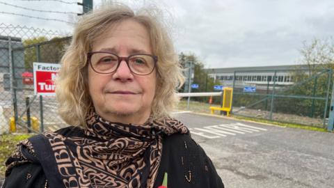 A woman with wavy blonde hair is looking at the camera. She is wearing dark brown glasses and a patterned black and brown scarf. Behind her is an industrial site.