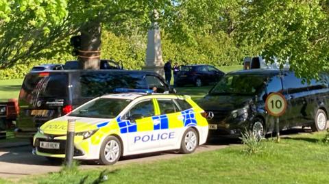 A police car parked on a road surrounded by grass and trees, with black vans and cars around it and a 10mph speed sign to the right.