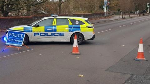 Street view of Riverside Road- with a white and yellow police car vertically placed on the road with a police sign in front of it and orange cones closing the road 