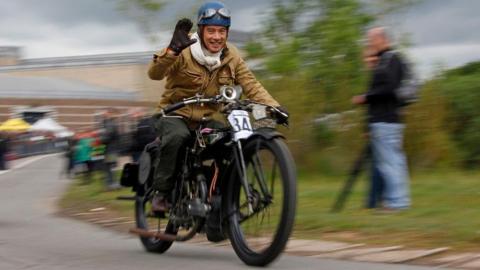 A man wearing a blue crash helmet with motorcycle goggles on it is riding a vintage motorcycle along a road. He is wearing a light brown bomber jacket and is smiling at the camera and waving a gloved hand.