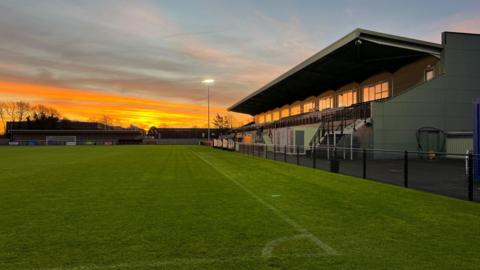 An image of Cambridge City's new stadium, showing the main stand with a sunrise scene in the background