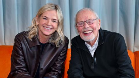 A woman and a mile sit side-by-side on an orange sofa smiling at the camera.