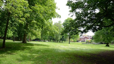 A green park with green grass and green trees. There is a house in the background with white windows.